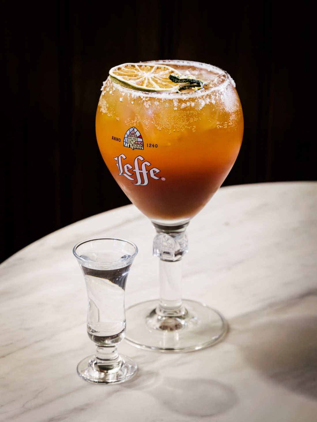 An amber cocktail with a dried lime wheel beside a shot glass on a marble table